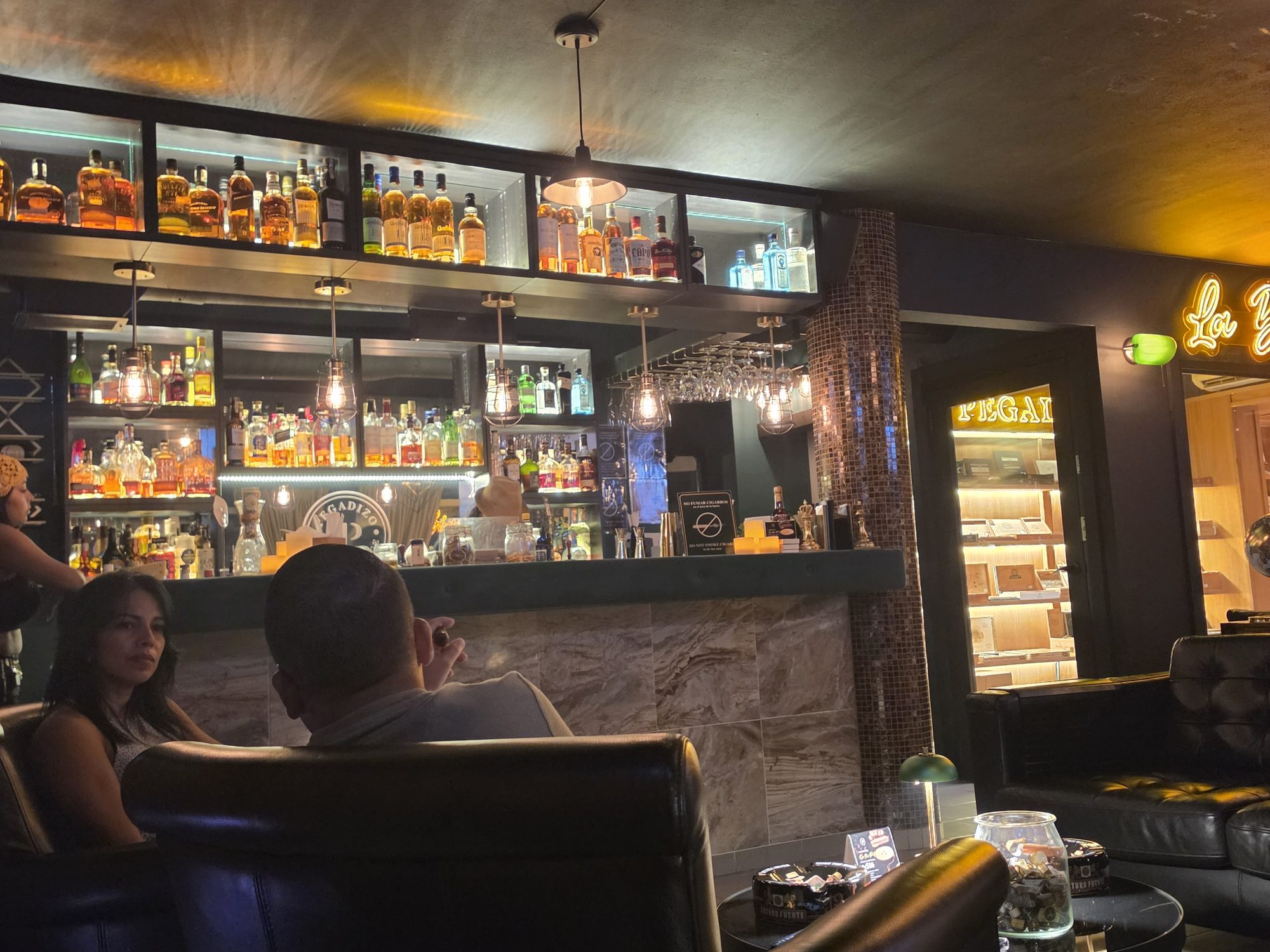 La barra de Pegadizo de noche, con la estantería de botellas iluminada y el humidor al fondo